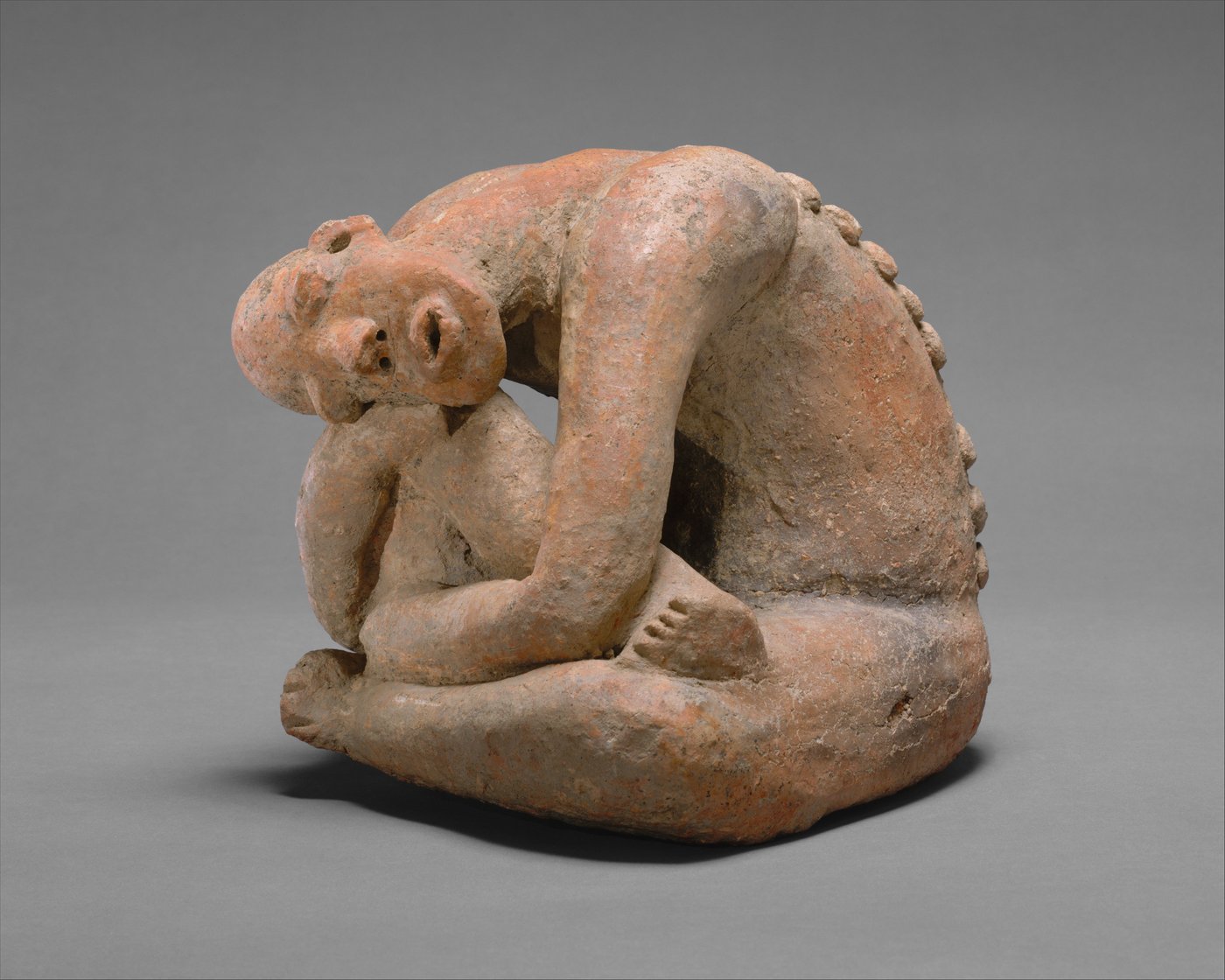 A seated terracotta figure with its arms wrapped around its raised knees