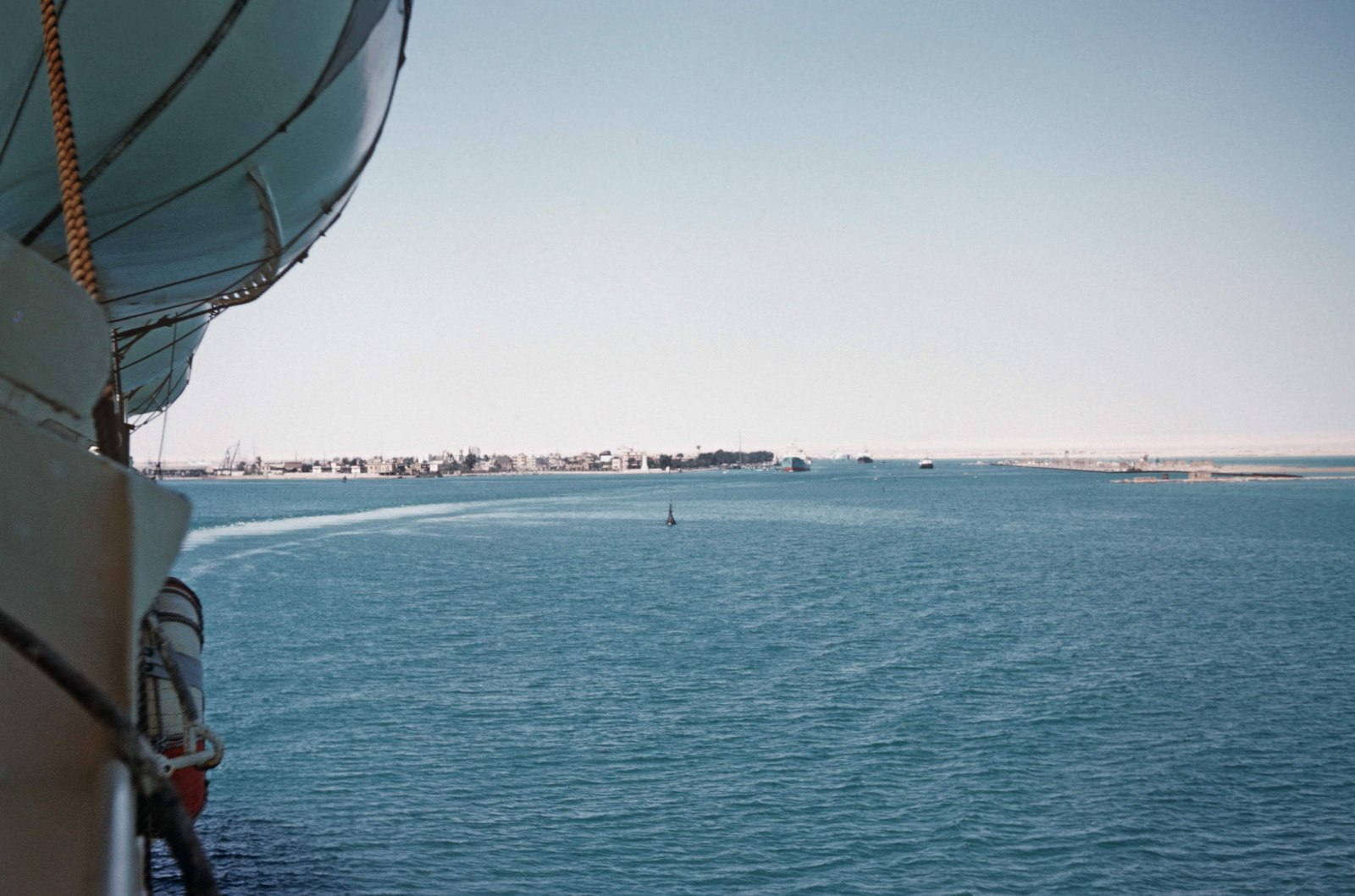 Vista desde un barco en 1965 hacia la entrada del canal de Suez, con aguas azules, edificios y otras embarcaciones a lo lejos.